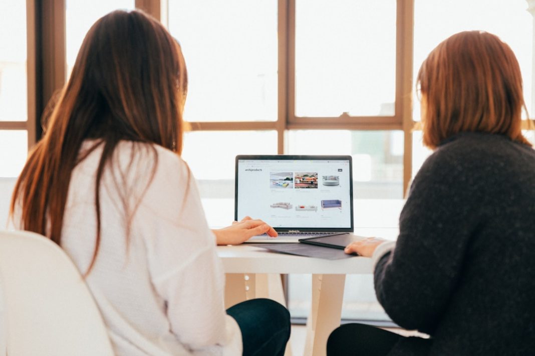 Two businesswomen sharing laptop to review sales coaching guide